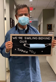 Doctor Lee holding a sign reading we are smiling behind our facemasks thank you
